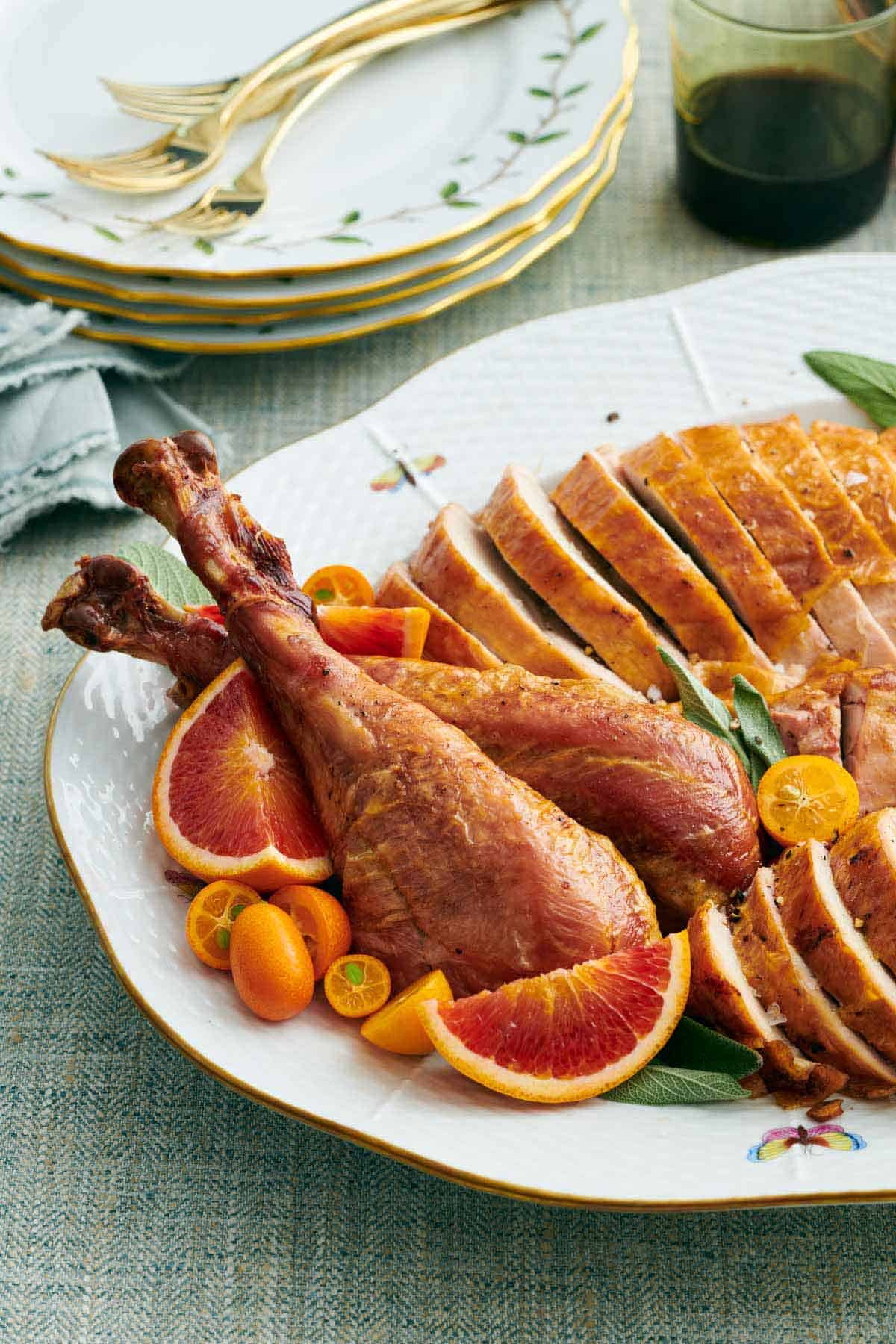 A close up of turkey legs surrounded by cut fruit along with the rest of the carved turkey. Stack of plates and forks in the background.