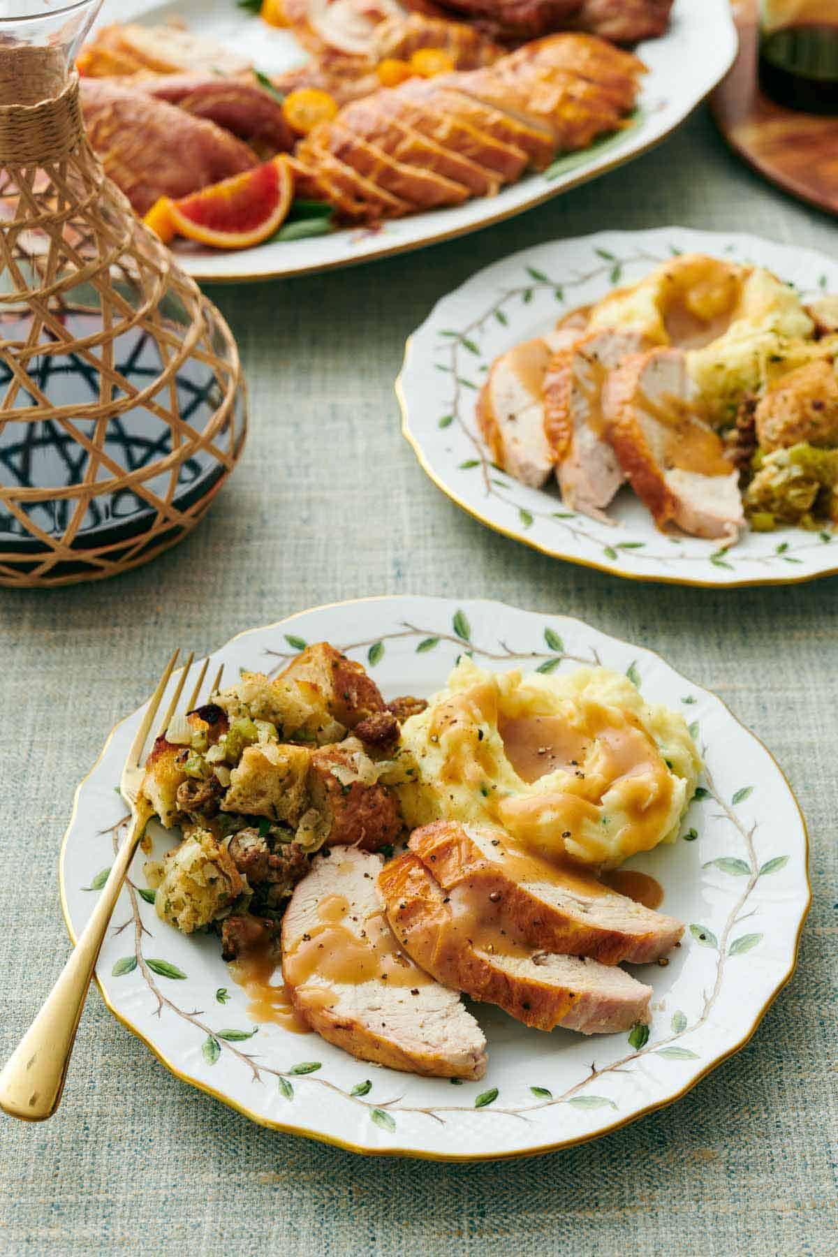 A plate with sliced turkey breasts, mashed potatoes with gravy, and stuffing. Another plated serving in the back along with the platter of carved turkey.