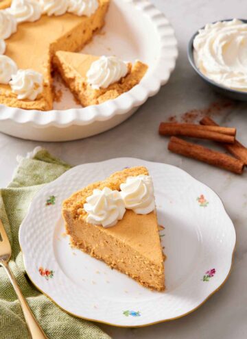 A slice of no-bake pumpkin cheesecake on a plate with a baking dish with the rest of the cheesecake in the background. Cinnamon sticks and a bowl of whipped cream on the side.