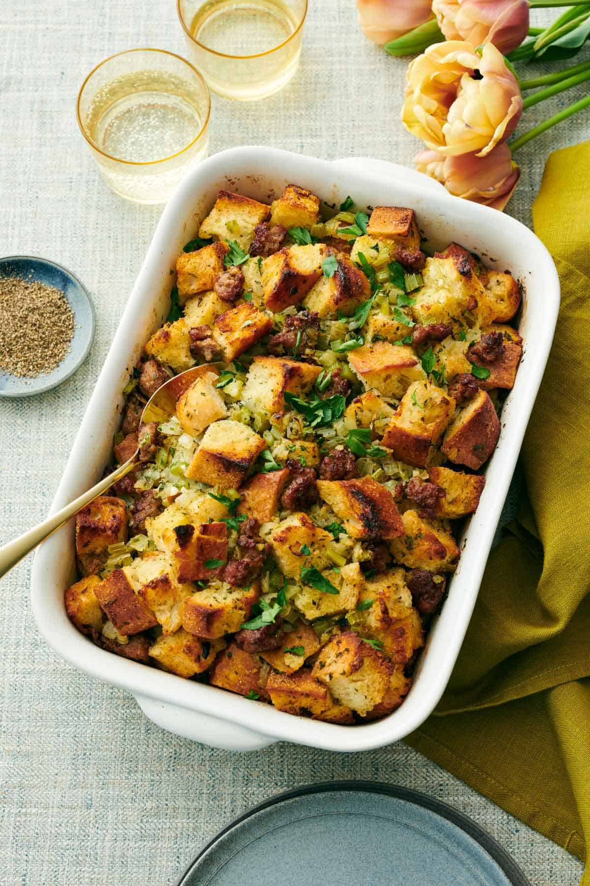 Overhead view of a white baking dish of sausage stuffing with a spoon inside. Flowers, two drinks, and a bowl of pepper on the side.