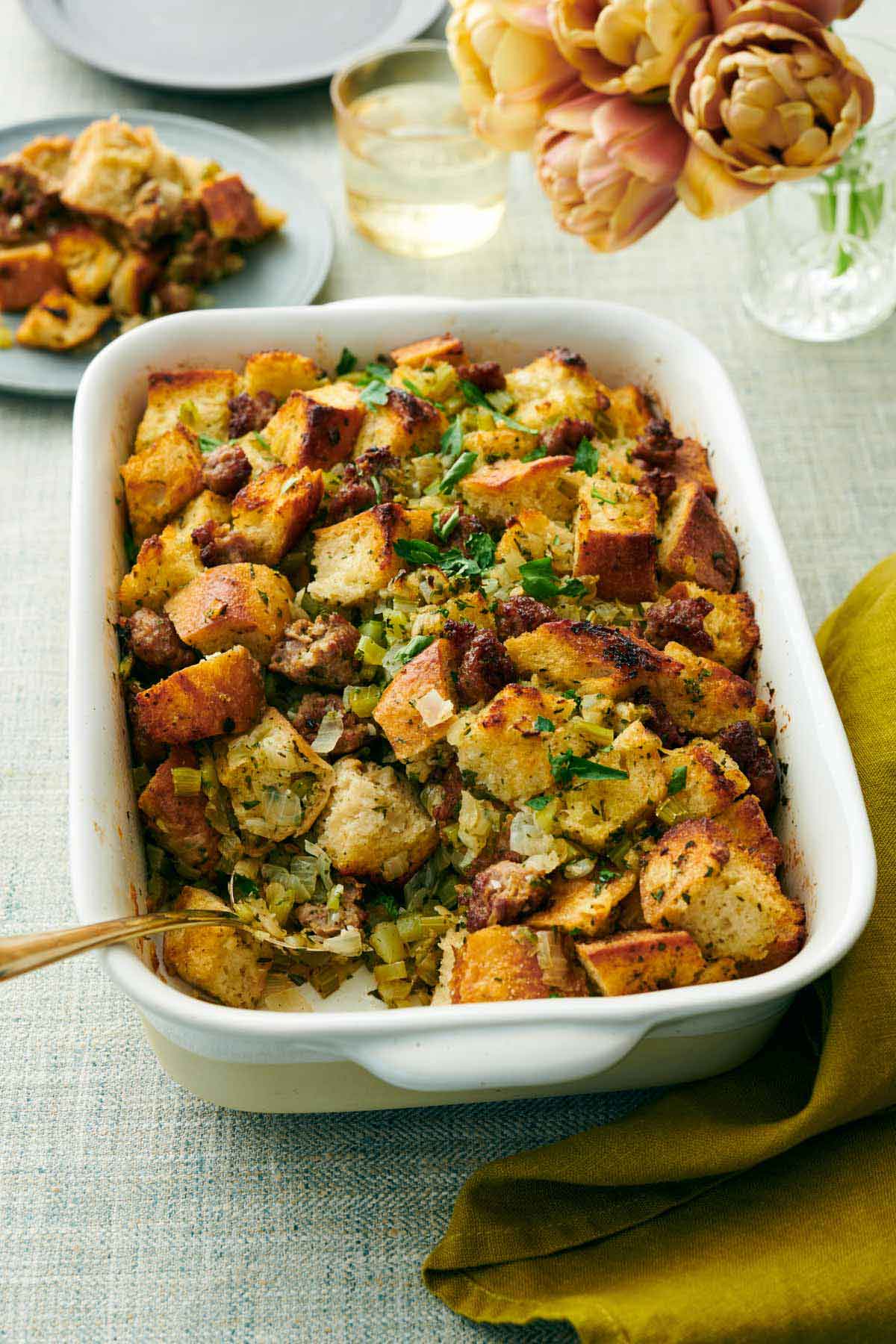 An angled view of a baking dish of sausage stuffing with a bit scooped out onto a plate in the background and a spoon inside. Flowers and drink in the back.