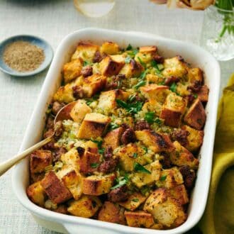 Pinterest graphic of a baking dish of sausage stuffing with a spoon inside. Flowers, drinks, bowl of pepper, and plates in the back.