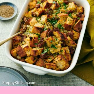 Pinterest graphic of a white baking dish of sausage stuffing with a spoon inside. Pepper, drinks, and flowers on the side.