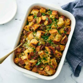 Overhead view of a white baking dish of sausage stuffing with a spoon inside. A linen napkin on the side and a plate.