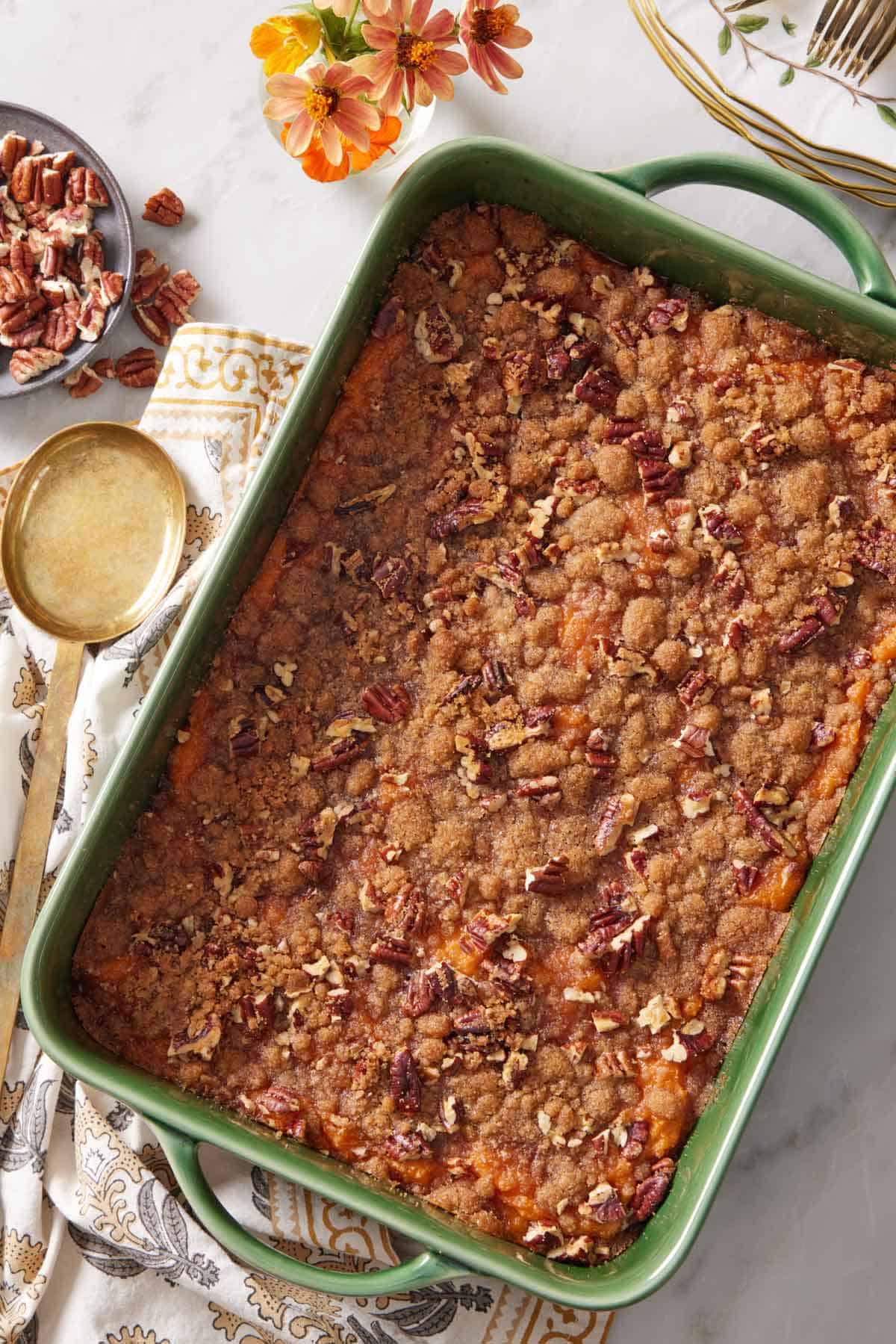 Overhead view of a green baking dish with a sweet potato casserole. A serving spoon, flowers, and pecans on the side.