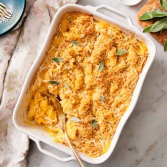 An overhead view of a baking dish of butternut squash mac and cheese with a serving spoon scooping some out. Fresh sage, a napkin, bowls, and forks on the side.