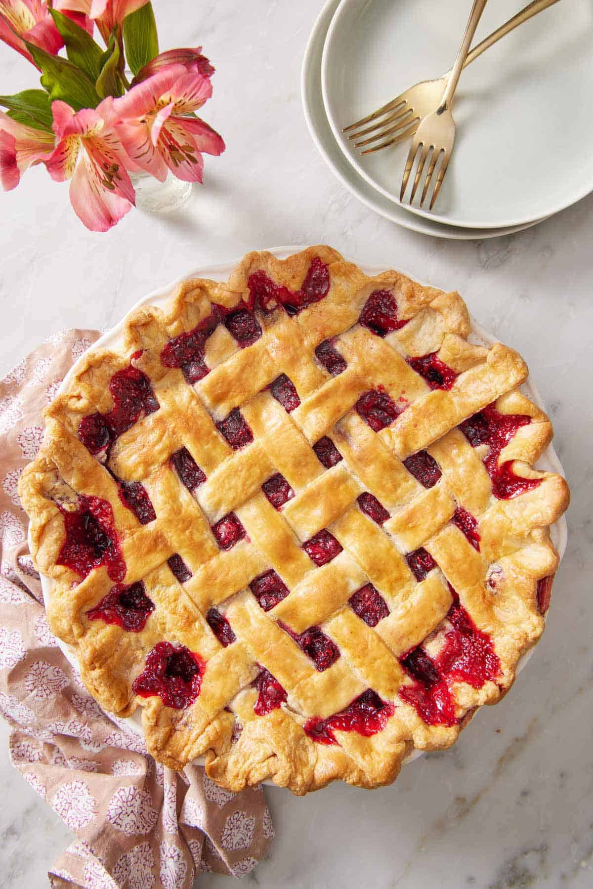 Overhead view of a cranberry pie with a lattice top crust. Flowers, a stack of platers, and forks off to the side.