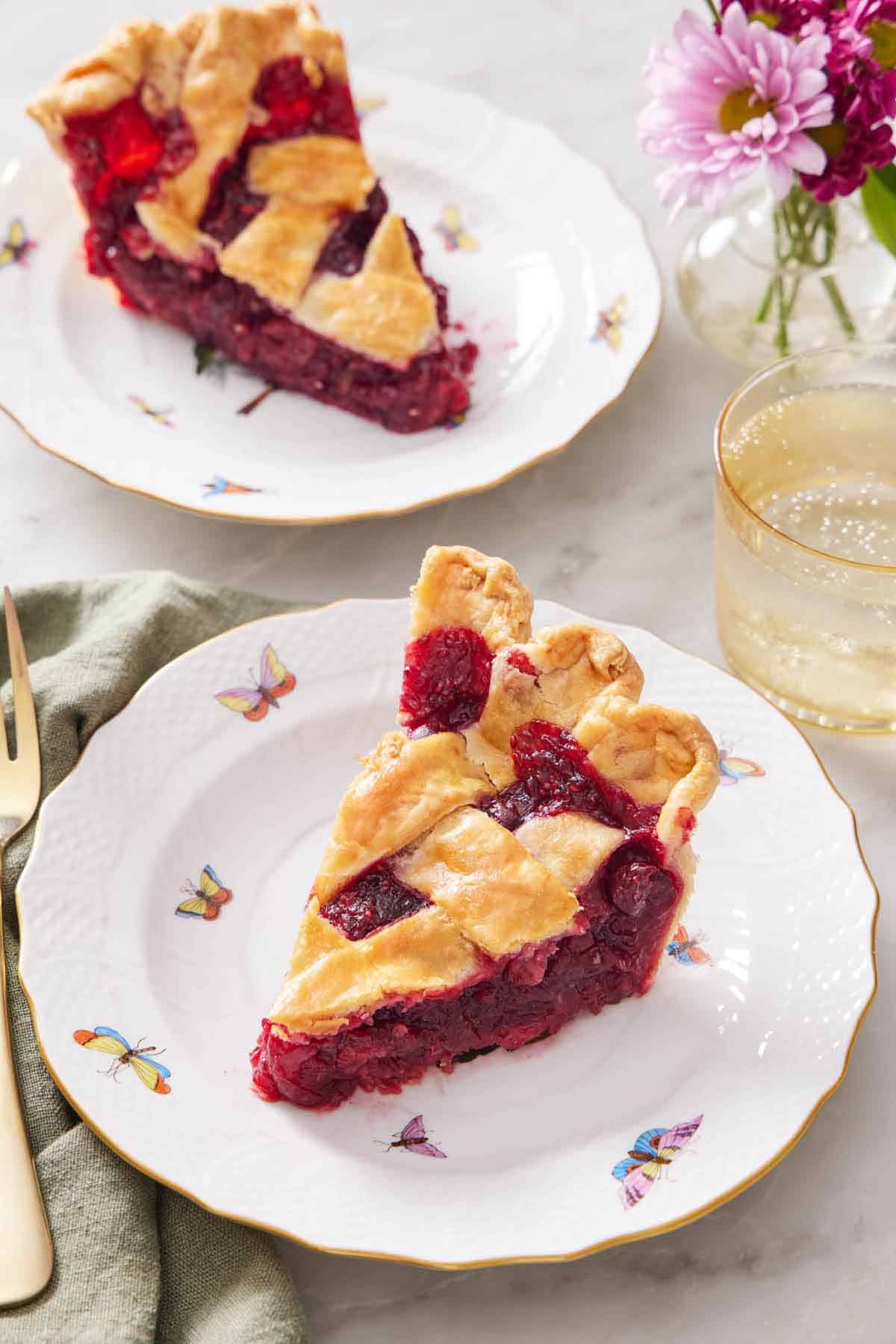 Two plates with a slice of cranberry pie with a glass of soda and vase of flowers off to the side.
