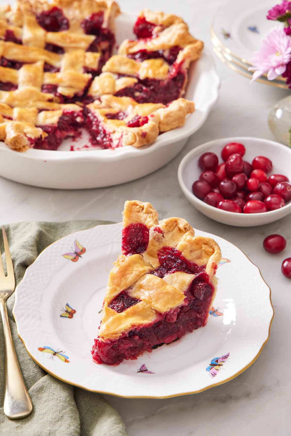 A slice of cranberry pie on a plate with a bowl of fresh cranberries and the rest of the pie in the background.