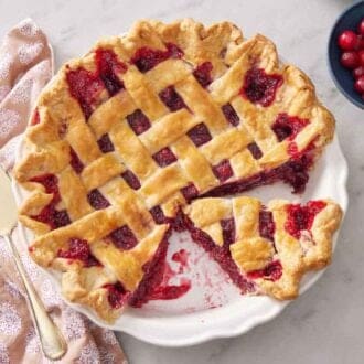 An overhead view of a cranberry pie in a backing dish with a few slices cut out and removed. A bowl of cranberries off to the side.