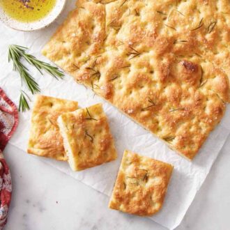 Overhead view of a slab of focaccia with three pieces cut out. Rosemary, a linen napkin, and a bowl of dipping oil on the side.