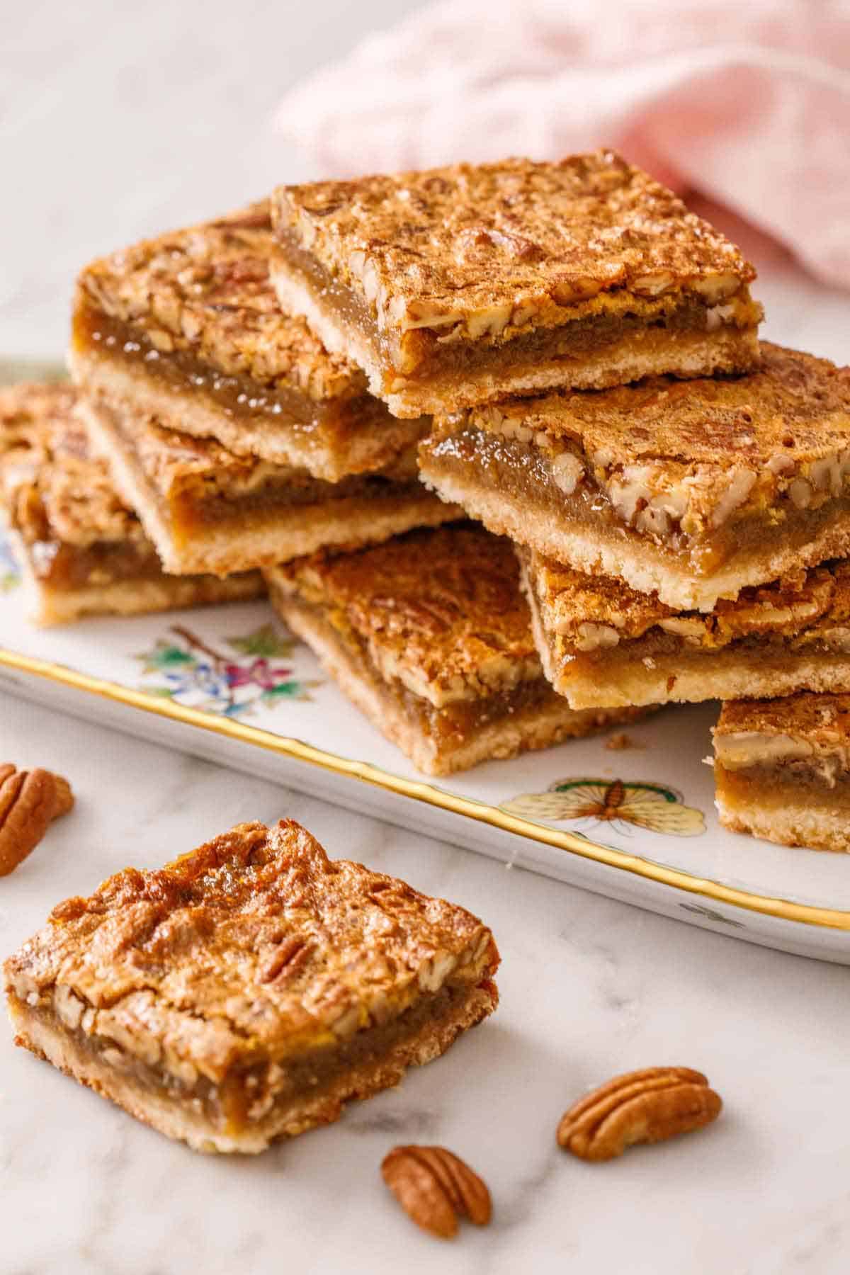 A platter with a stack of pecan pie bars with one bar in front on the counter with some pecans scattered around.