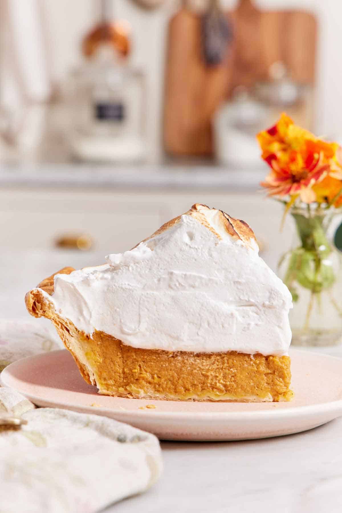 A profile view of a slice of pumpkin meringue pie, showing off its height, on a plate with flowers in the background.