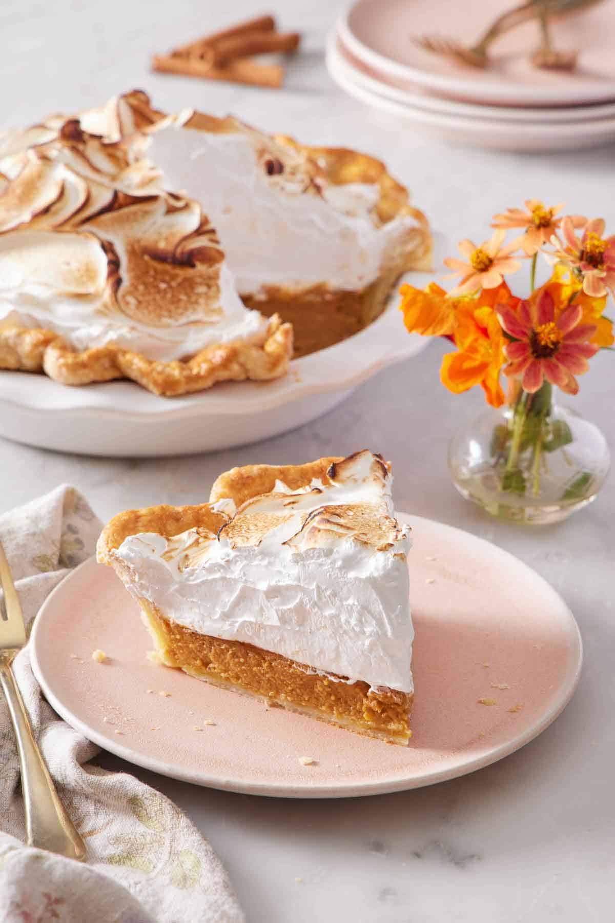 A plate with a slice of pumpkin meringue pie with flowers in the background along with the rest of the pie.