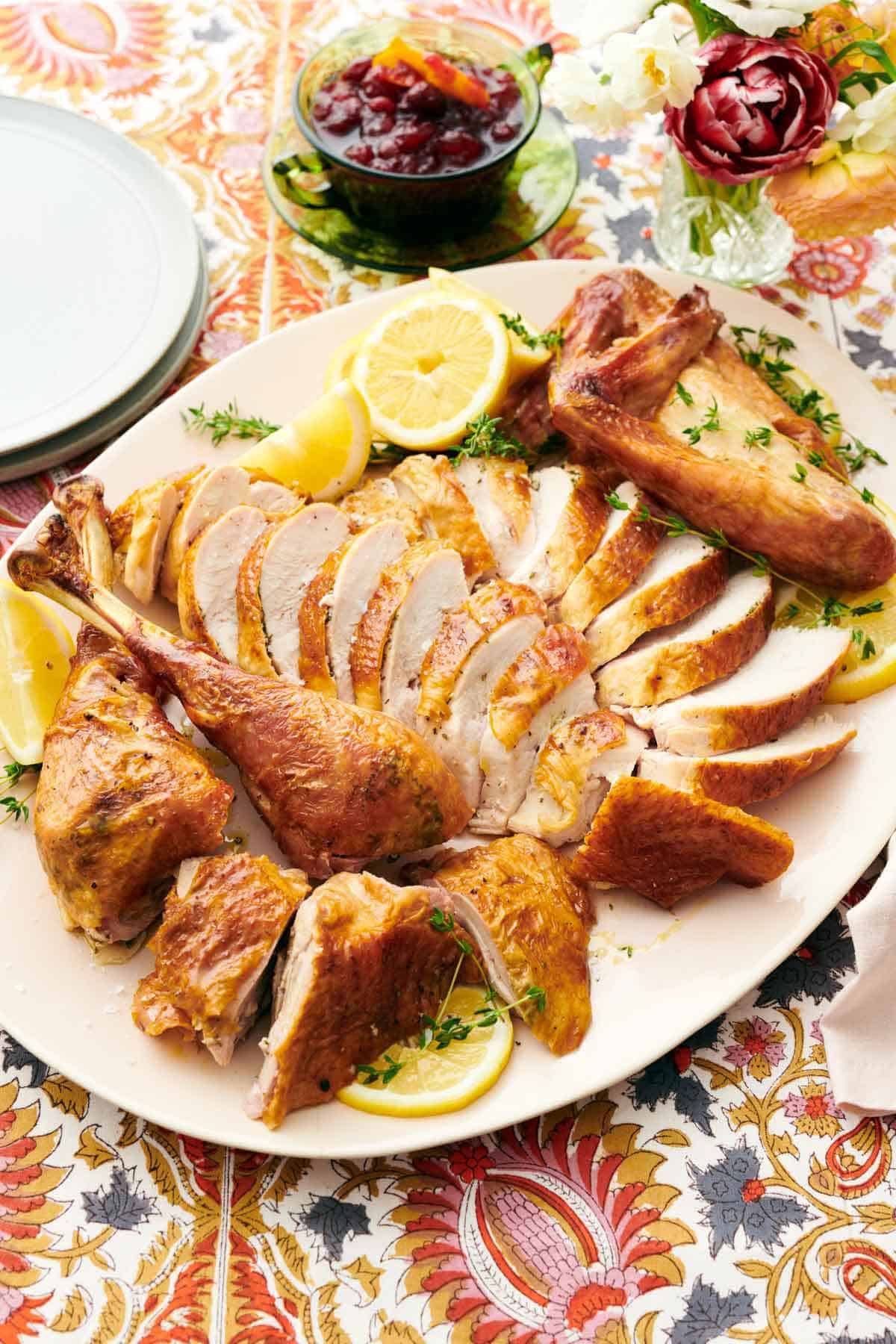 A platter with a sliced spatchcock turkey surrounded by fresh herbs and cut lemons on a floral tablecloth. Flowers, cranberry sauce, and a stack of plates on the side.