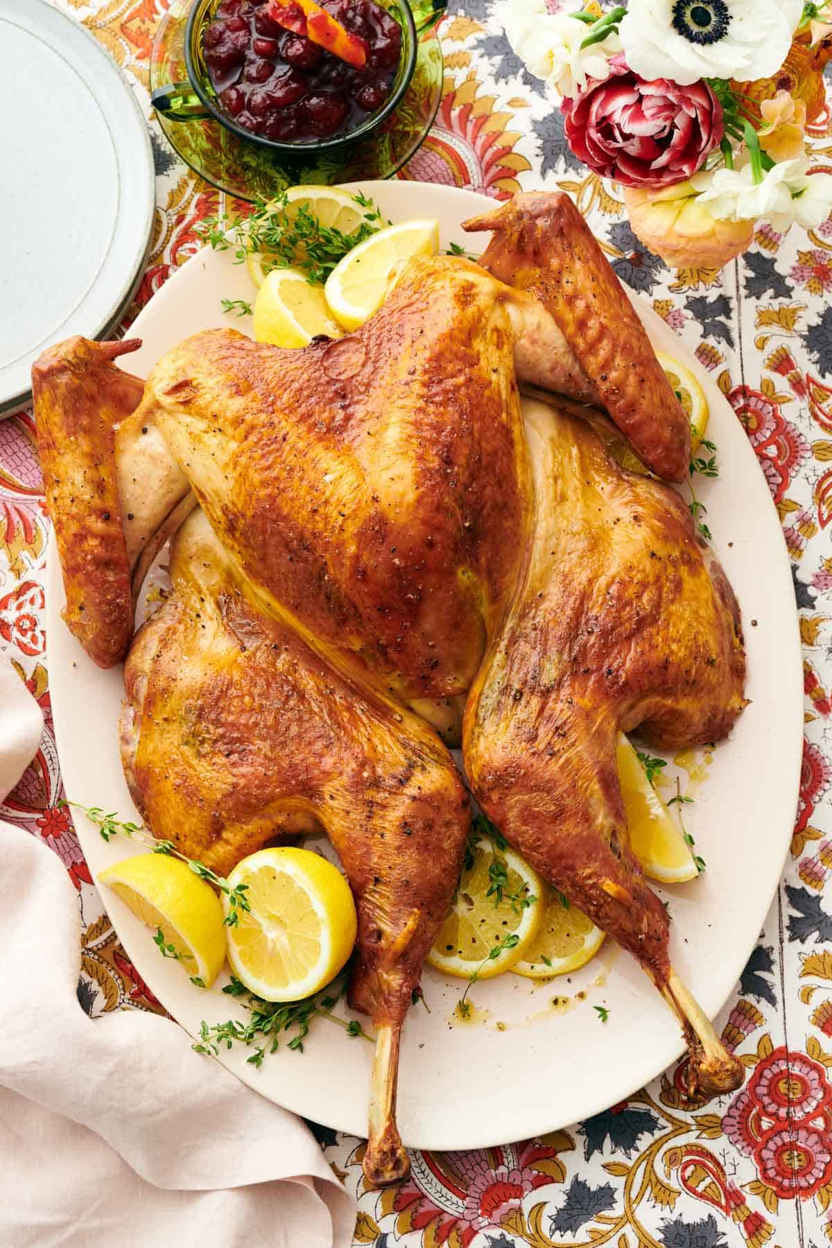 Overhead view of a platter with a spatchcock turkey surrounded by fresh herbs and lemons. A bowl of cranberry sauce and plates off to the side.