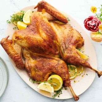 Overhead view of a platter of spatchcock turkey with cut lemons and fresh herbs. Flowers and a stack of plates on the side.