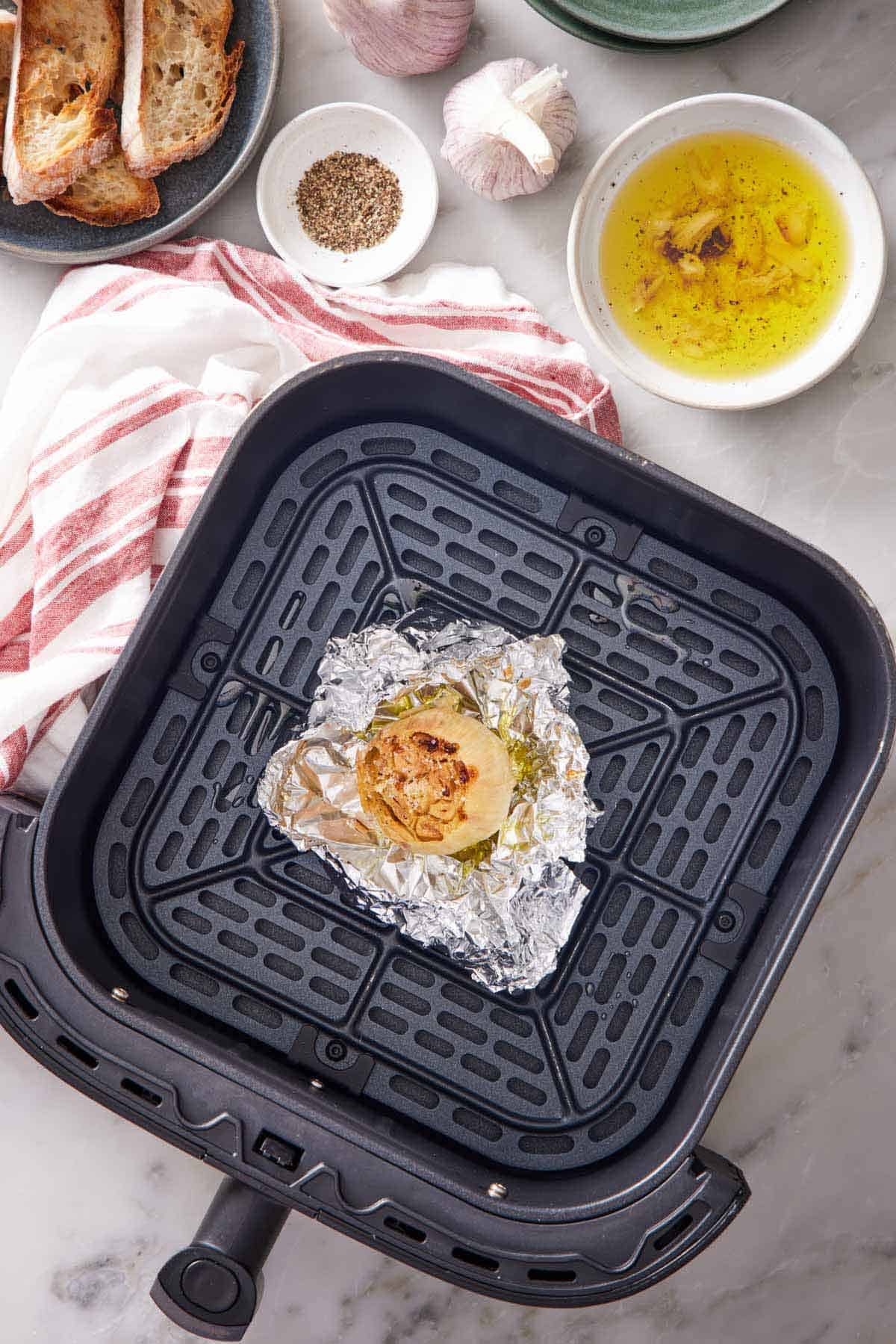 An overhead view of air fryer roasted garlic in foil in the air fryer basket. Dipping oil, pepper, garlic heads, and bread off to the side.