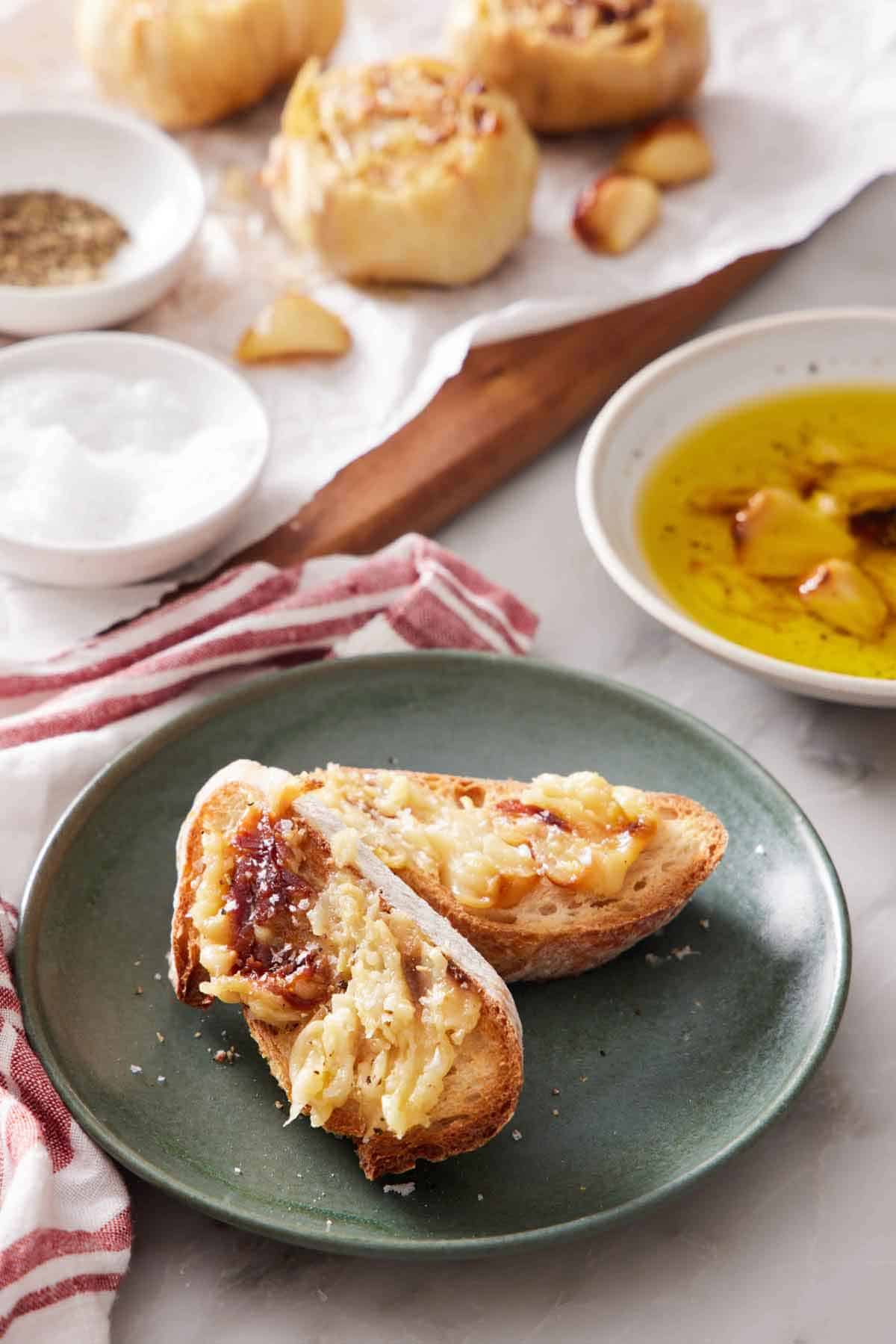 A plate with two pieces of bread with air fryer roasted garlic spread on top. Dipping oil in a bowl in the back along with more roasted heads and salt and pepper.