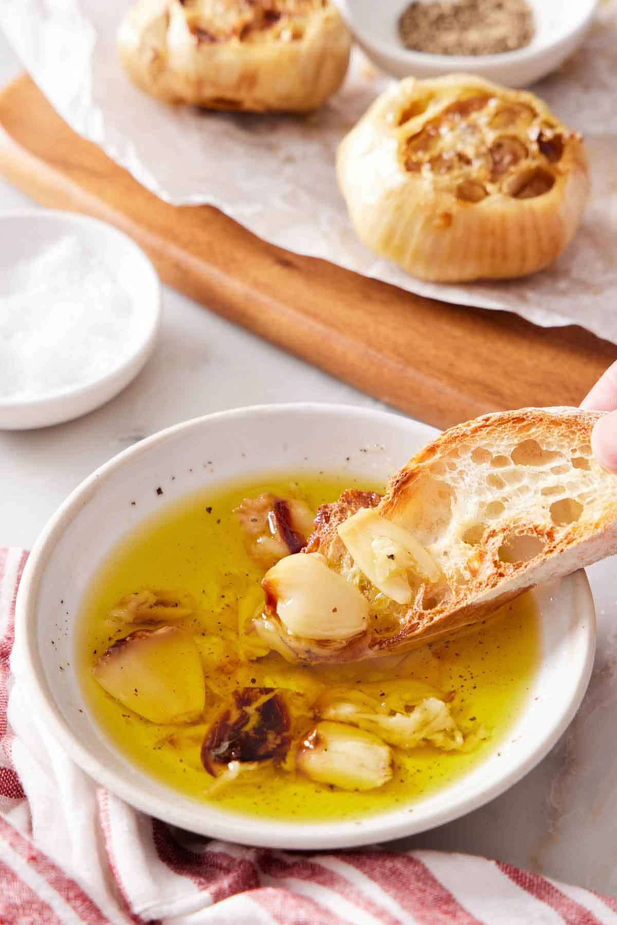 A piece of bread dipped into oil with air fryer roasted garlic. More roasted heads in the background.