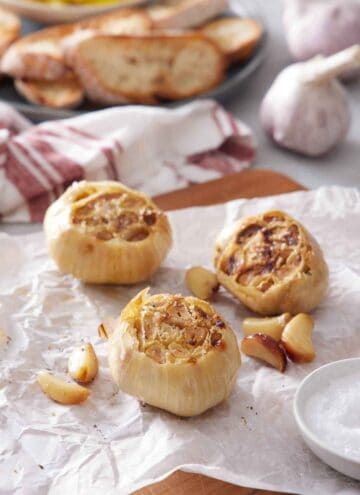 Three air fryer roasted garlic also with roasted cloves on a parchment-lined wooden serving board. More garlic and sliced bread in the background.