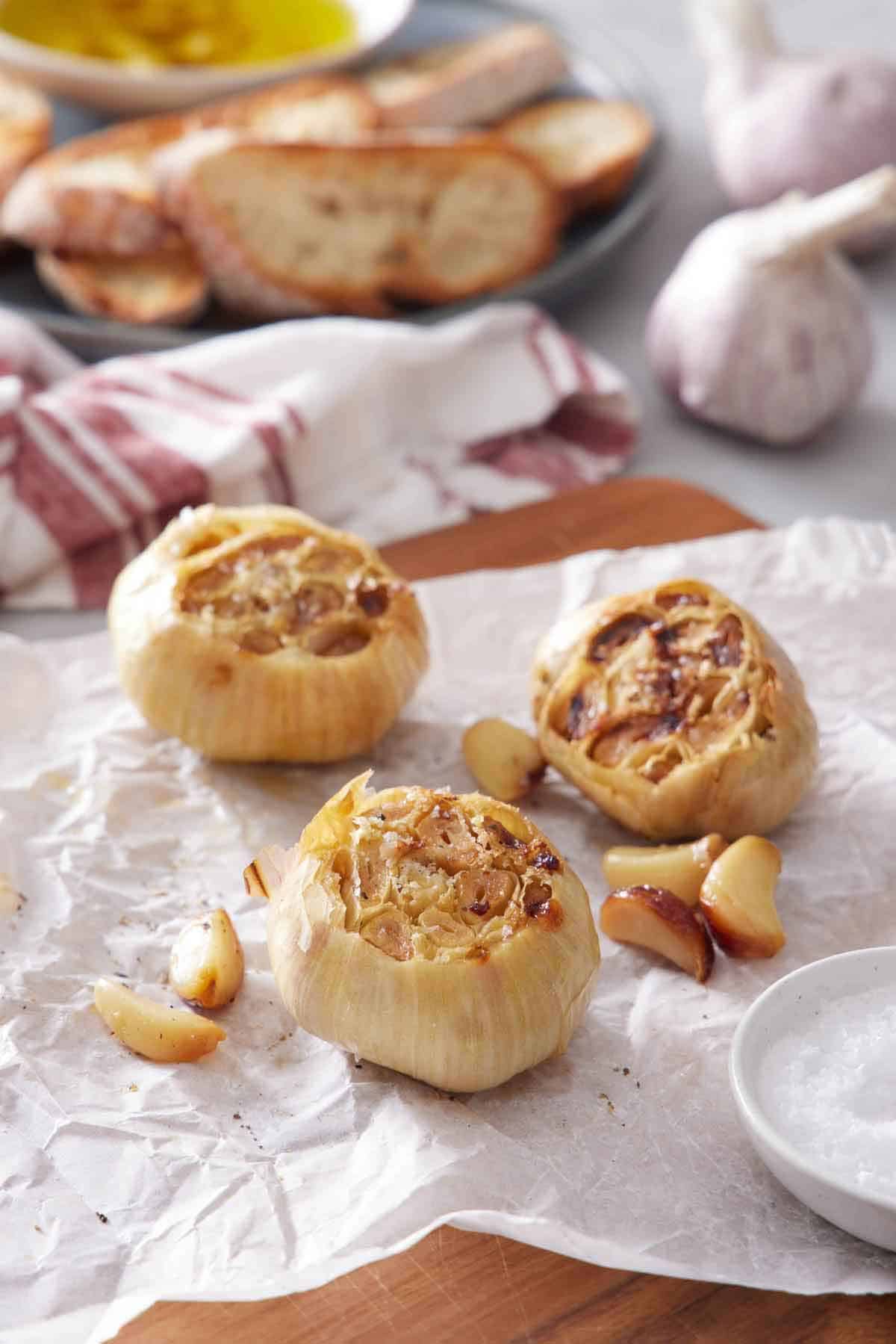 Three air fryer roasted garlic also with roasted cloves on a parchment-lined wooden serving board. More garlic and sliced bread in the background.