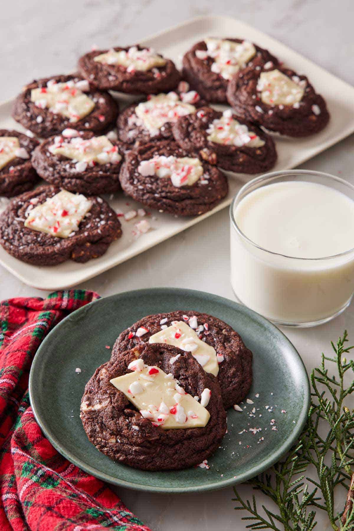 A plate with two chocolate peppermint bark cookies. A glass of milk and platter with more cookies in the background.