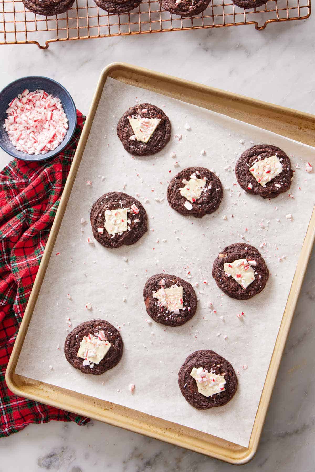 Overhead view of eight chocolate peppermint bark cookies on a parchment-lined sheet pan. A bowl of crushed candy canes on the side.