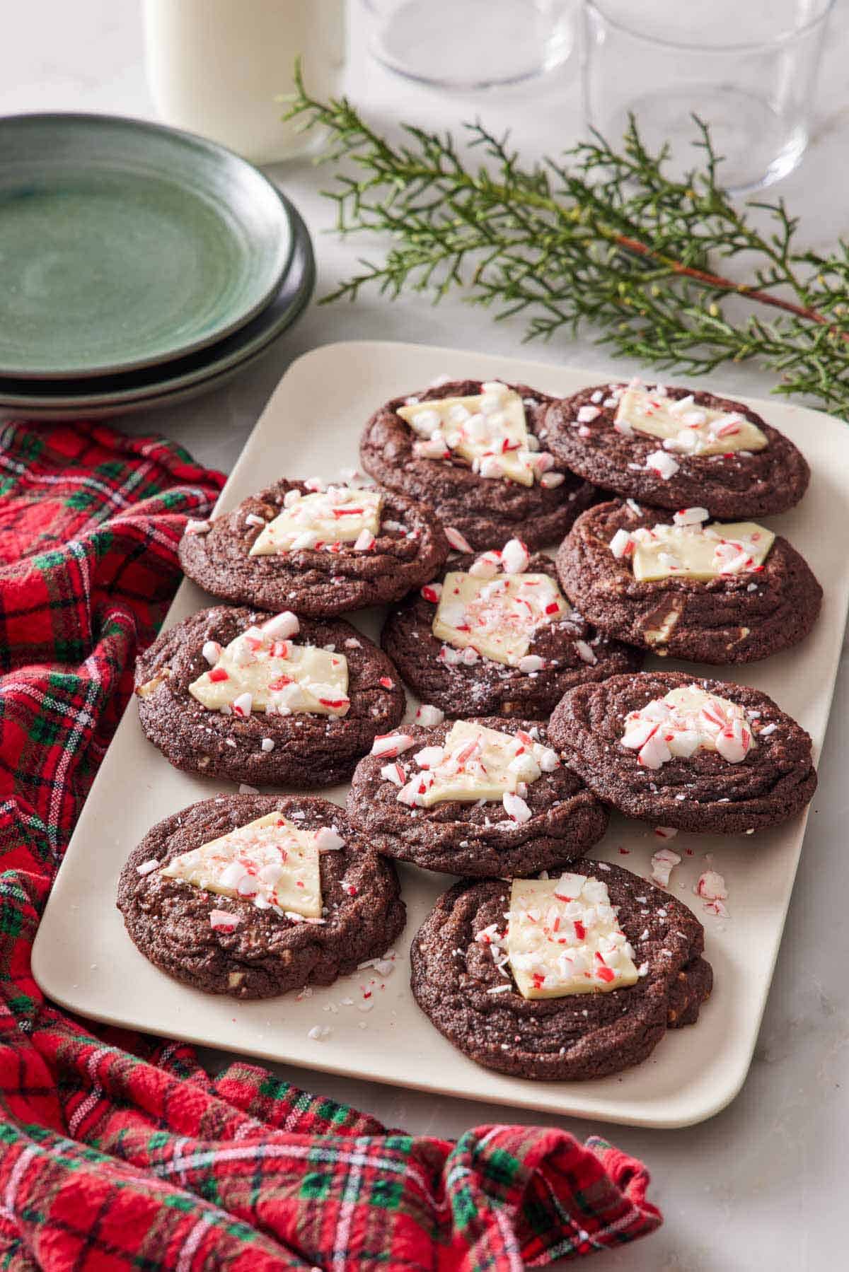 A platter of chocolate peppermint bark cookies. Decorative pines, plates, and a checkered linen napkin surrounding the platter.