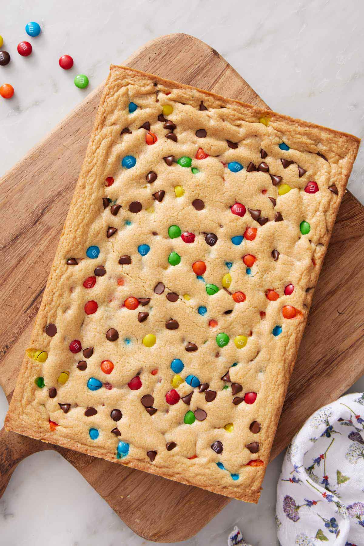 Overhead view of an uncut slab of M&M cookie bars on a wooden serving board. M&M's and a linen napkin on the side.