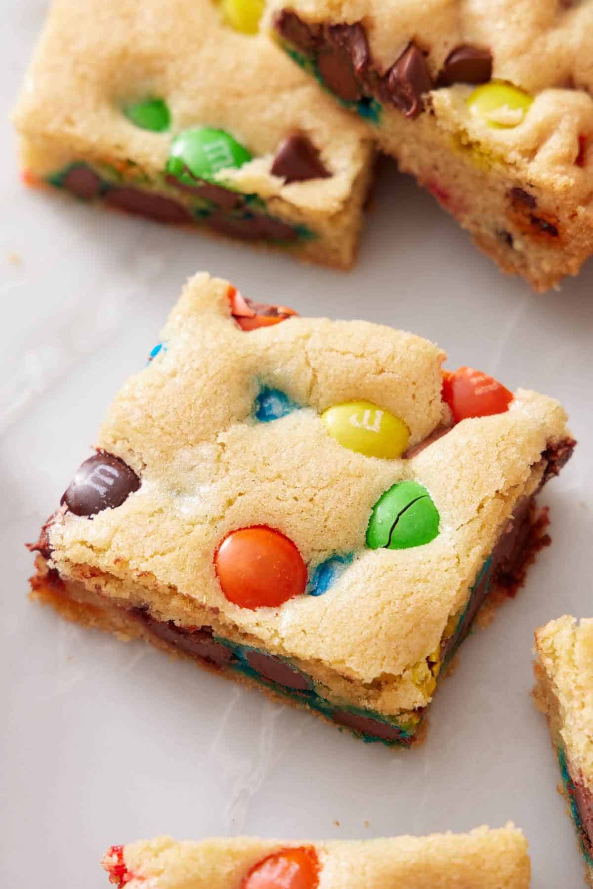 A piece of M&M cookie bar on a marble surface. More on the side.
