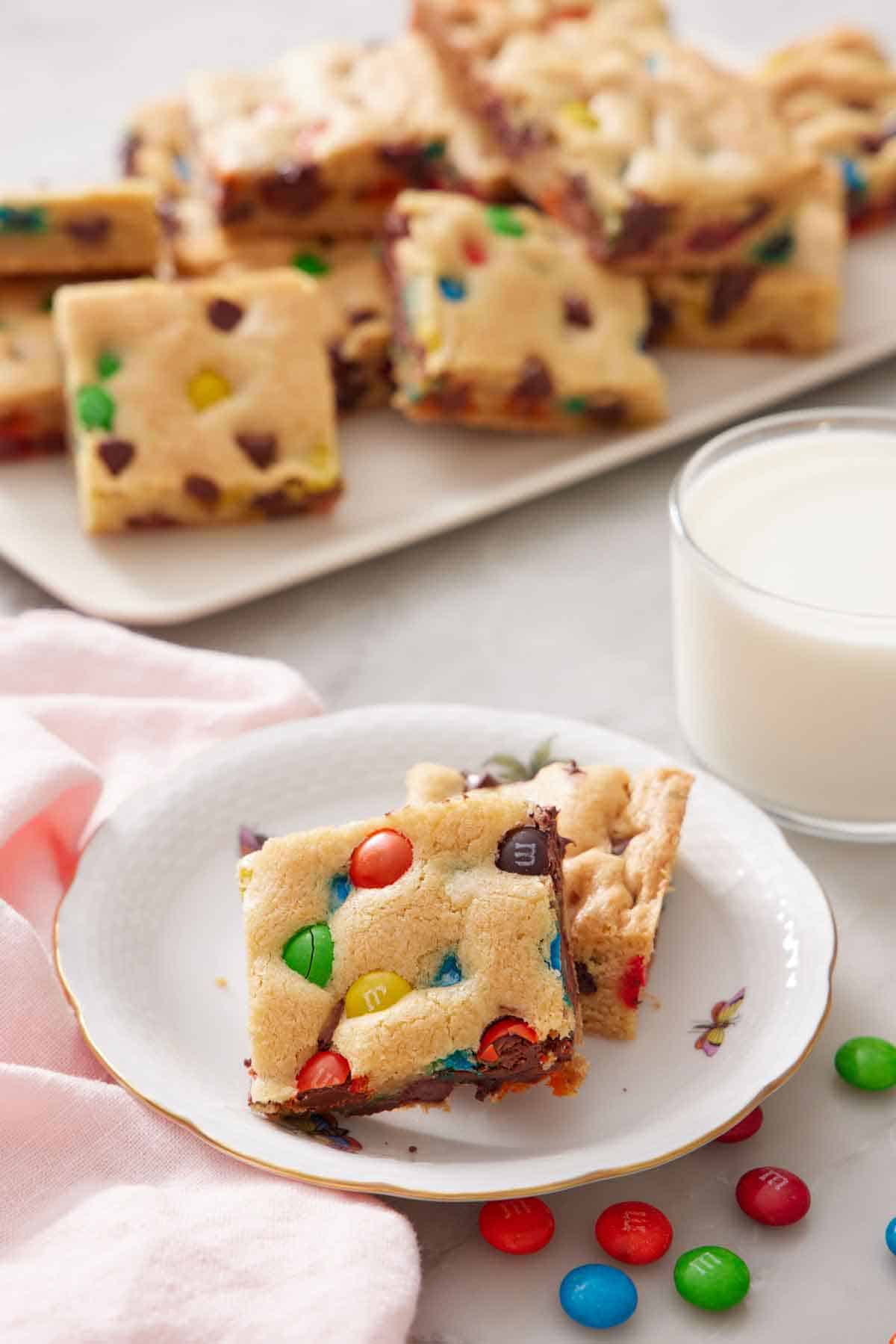 Two pieces of M&M cookie bars on a plate with a platter of more and a glass of milk in the background.