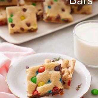 Pinterest graphic of two pieces of M&M cookie bars on a plate with a platter of more and a glass of milk in the background.
