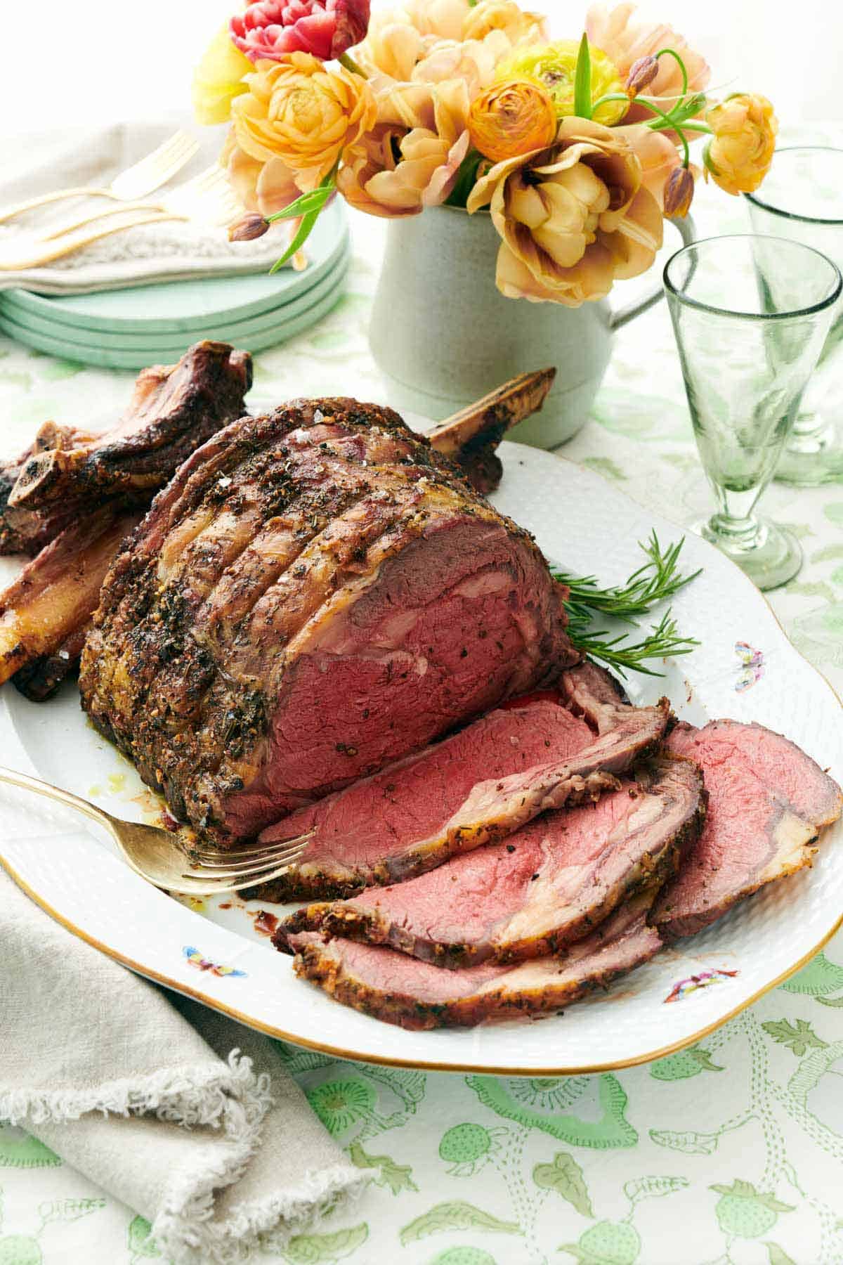 A platter with a prime rib with a few slices cut in front of it. A vase of flowers in the back along with some cups and plates.