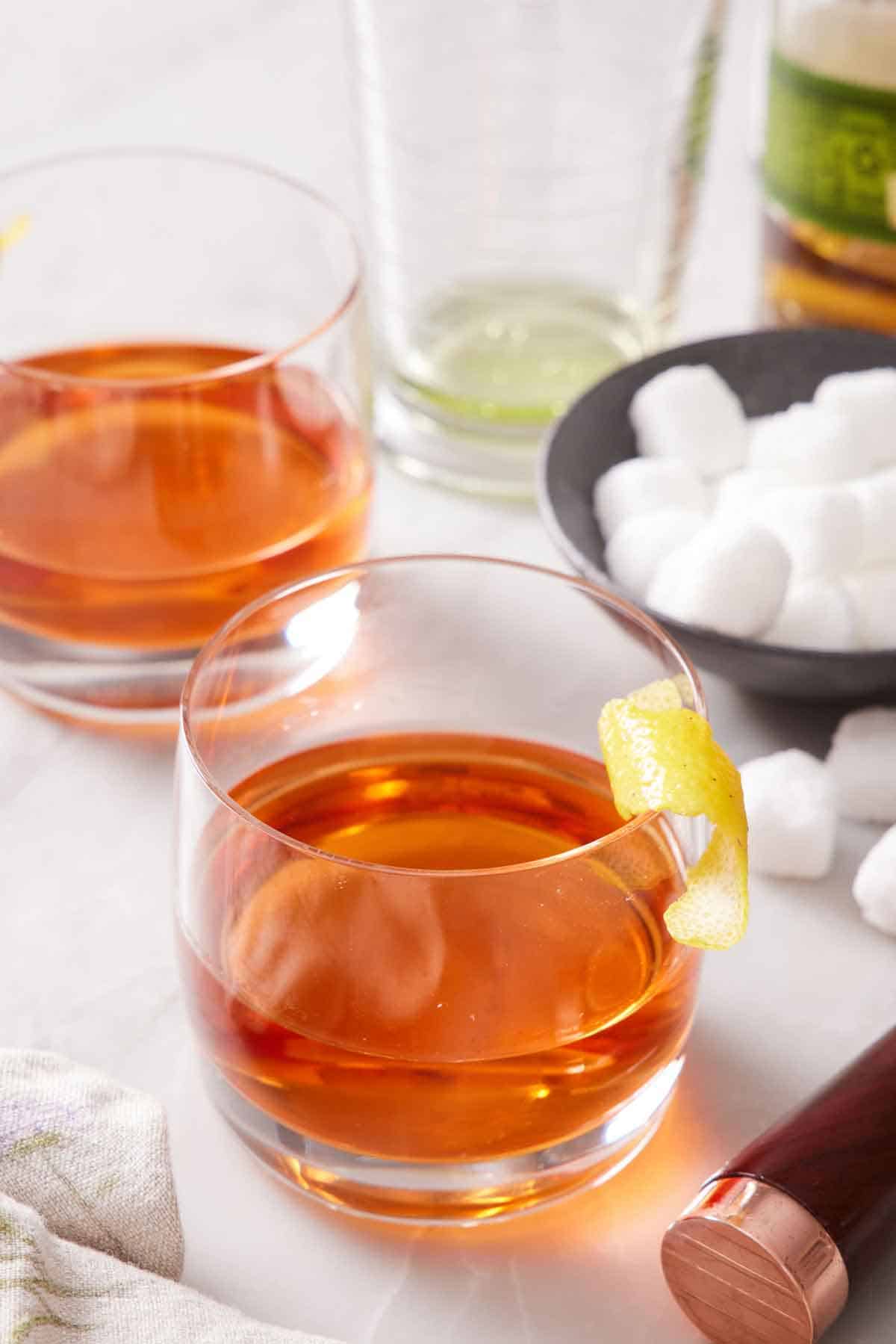 Two glasses of Sazerac with a lemon twist garnish. A bowl of sugar cubes in the back.