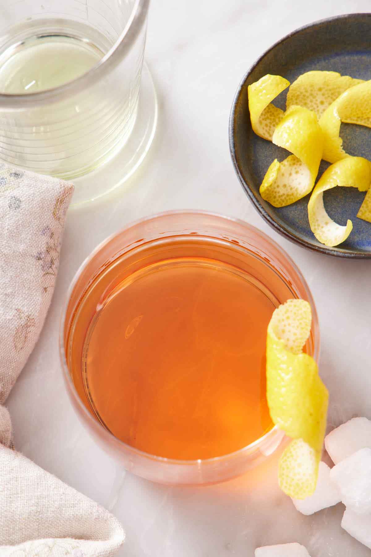 Overhead view of a glass of Sazerac with a lemon zest garnish with more garnishes in a bowl off to the side along with scattered sugar cubes.