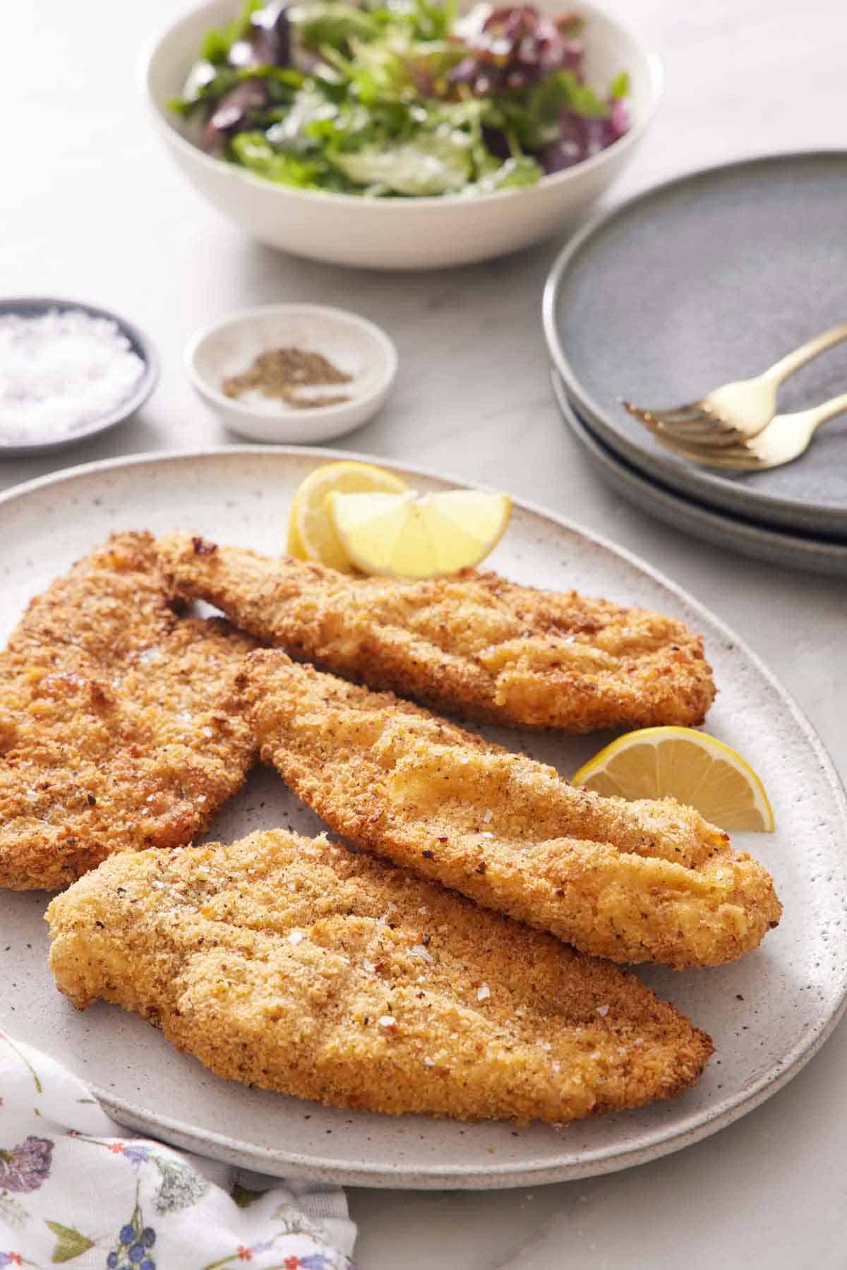 A platter of air fryer chicken cutlets with cut lemon wedges. A bowl of salad in the back along with salt, pepper, stack of plates, and forks.
