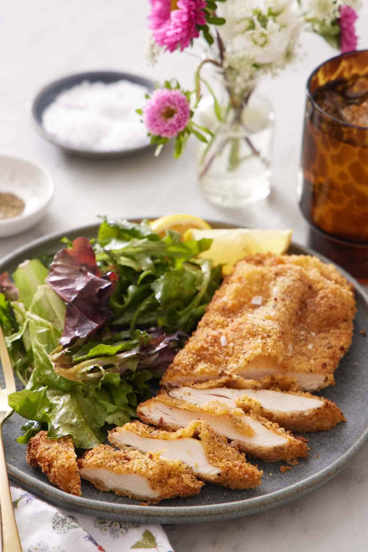 A plate with an air fryer chicken cutlet with half of it sliced along with some mixed greens. Flowers, a drink, salt, and pepper in the background.