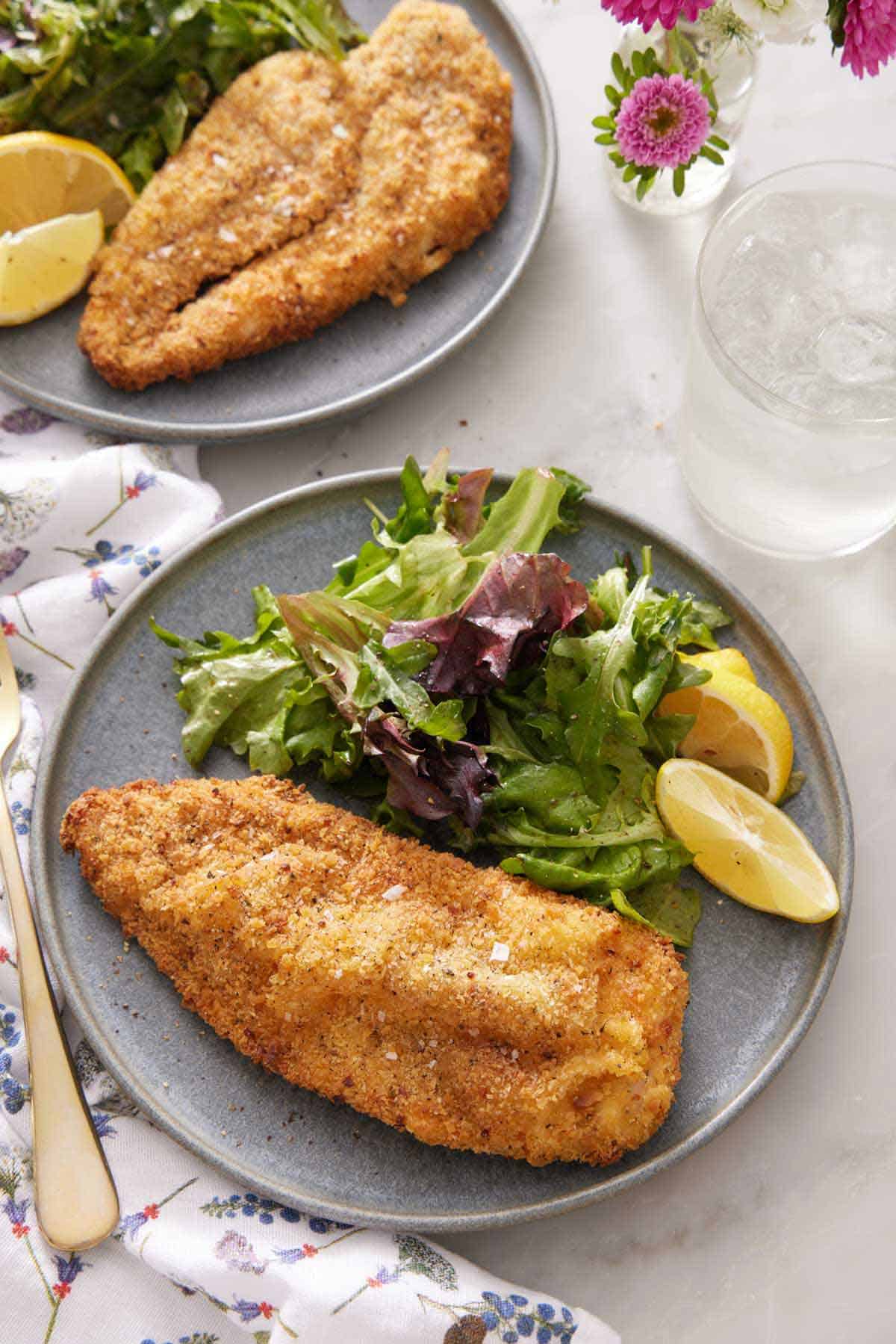 Overhead view of a plate with an air fryer chicken cutlet, mixed greens, and lemon wedges. A drink and another plated serving off to the side.
