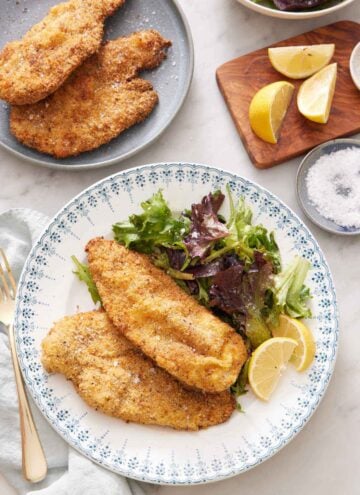 A plate with crispy air fryer chicken cutlets along with a side of mixed greens and lemon wedges. More cutlets in the back along with cut lemon wedges and a bowl of salt.