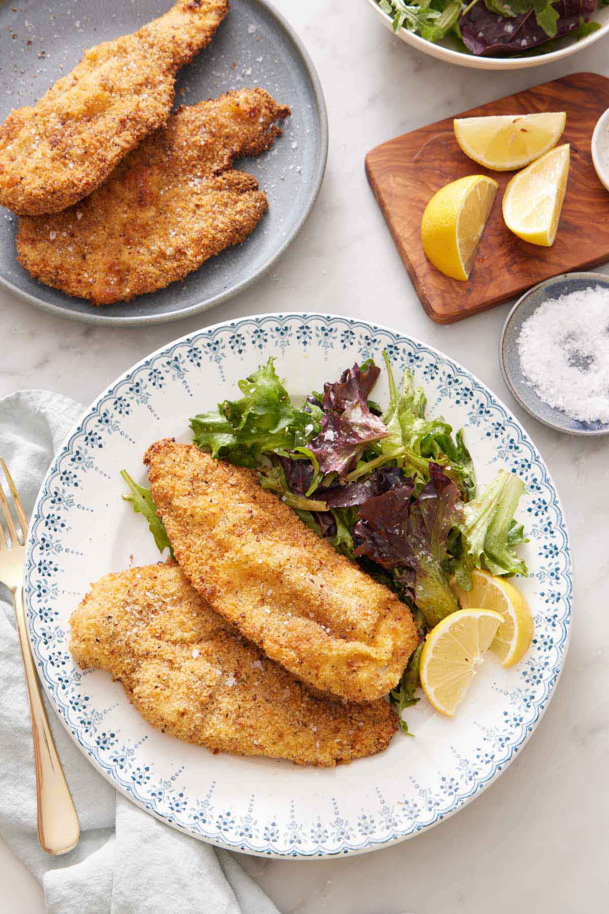 A plate with crispy air fryer chicken cutlets along with a side of mixed greens and lemon wedges. More cutlets in the back along with cut lemon wedges and a bowl of salt.