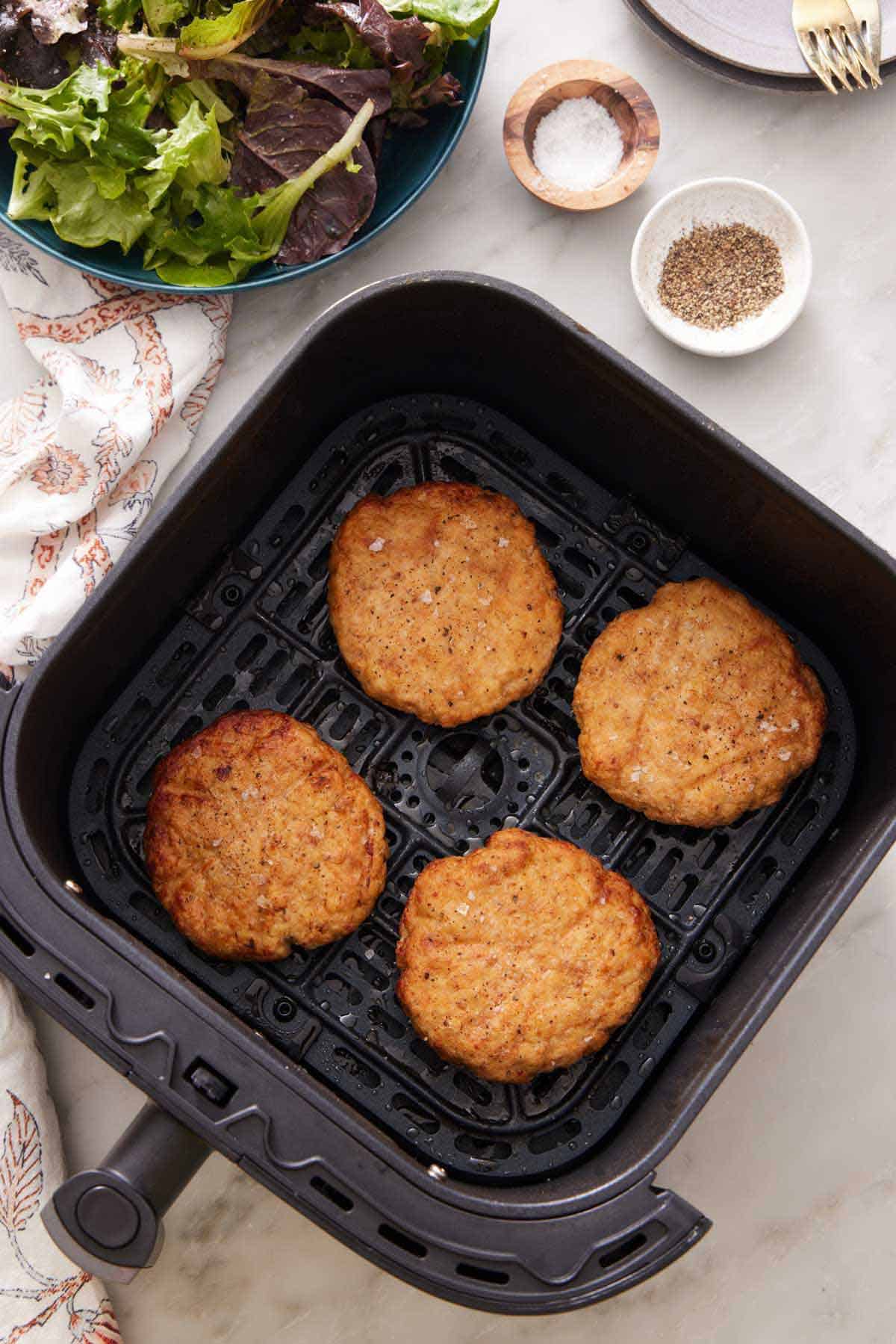 Overhead view of an air fryer basket with four air fryer chicken patties. Salt, pepper, and lettuce on the side.
