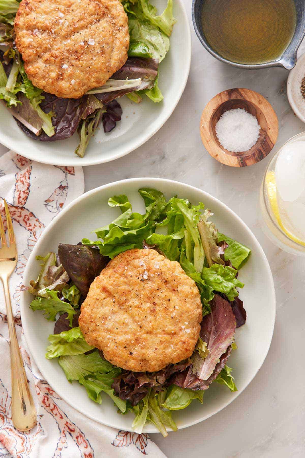 Overhead view of two plates of lettuce with an air fryer chicken patty on top. Salt and a bowl of dressing on the side.