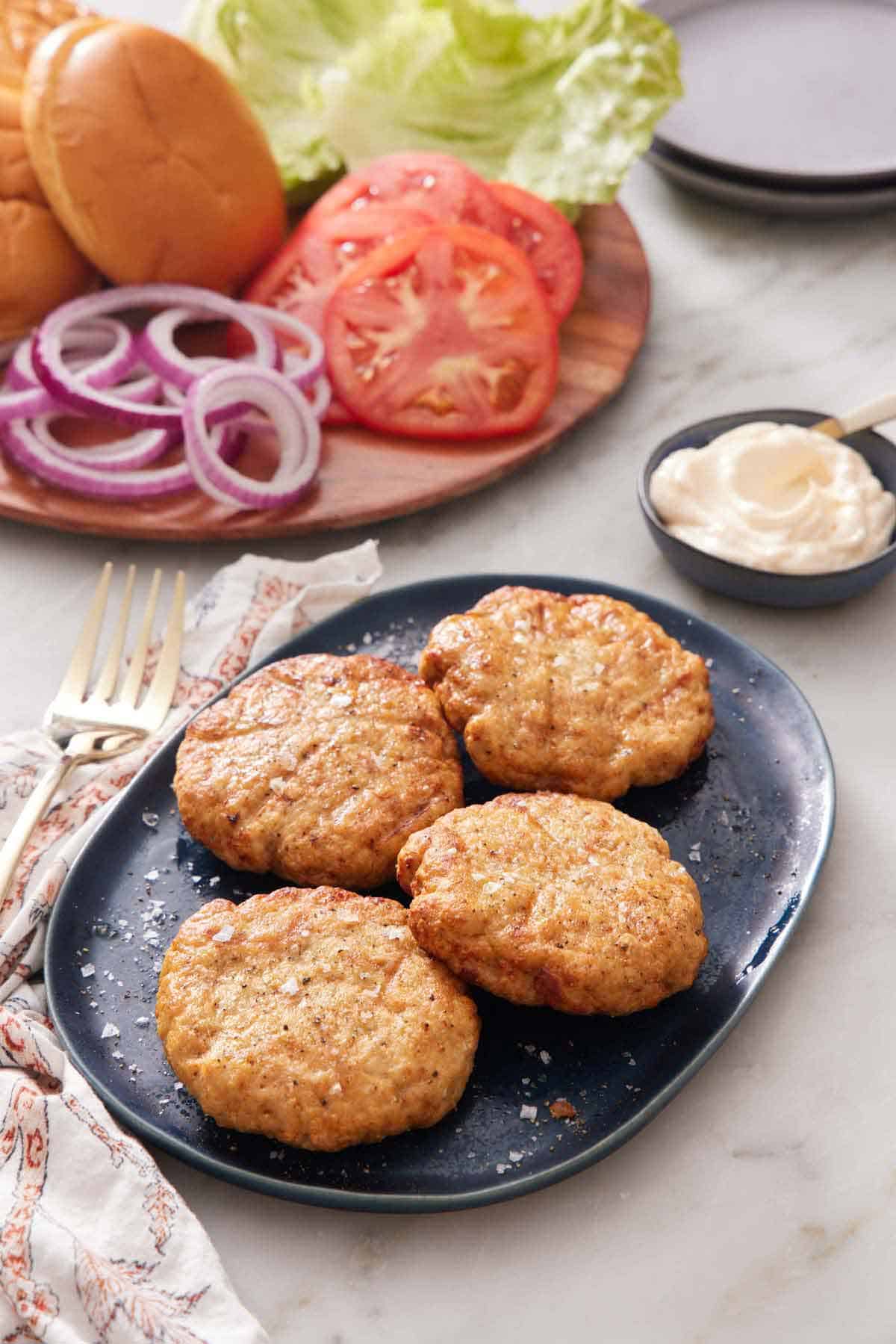 A platter with four air fryer chicken patties. Burger toppings in the back along with a bowl of sauce.