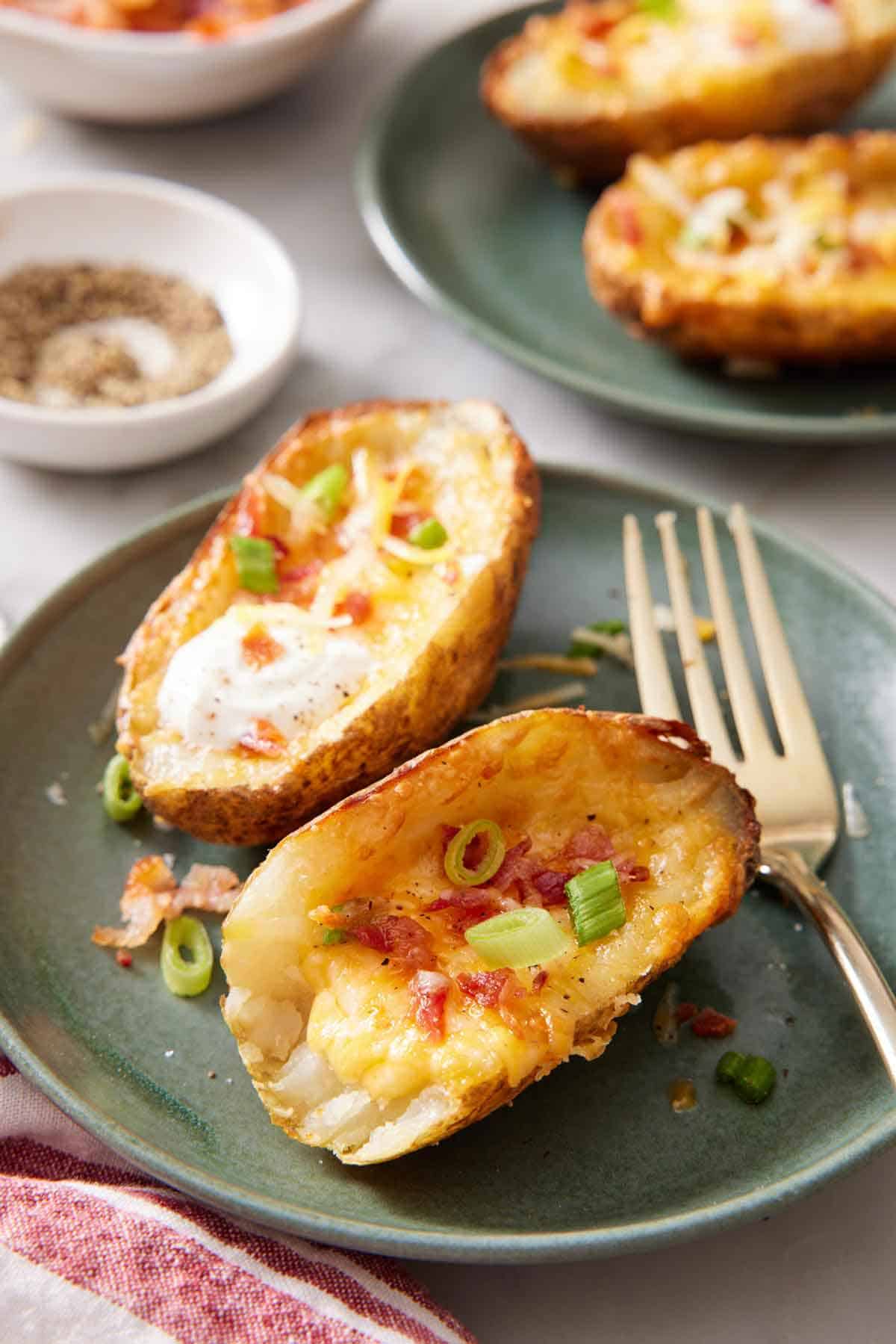 A green plate with two air fryer potato skins with green onions on top with a fork on the side. Pepper and more air fryer potato skins in the background.