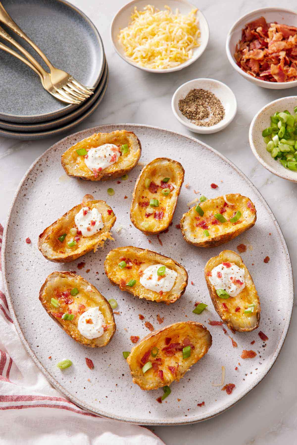 An overhead view of a platter of air fryer potato skins with green onion garnished on top. A few potato skins with dollops of sour cream on top. A stack of plates and bowls of toppings off to the side.