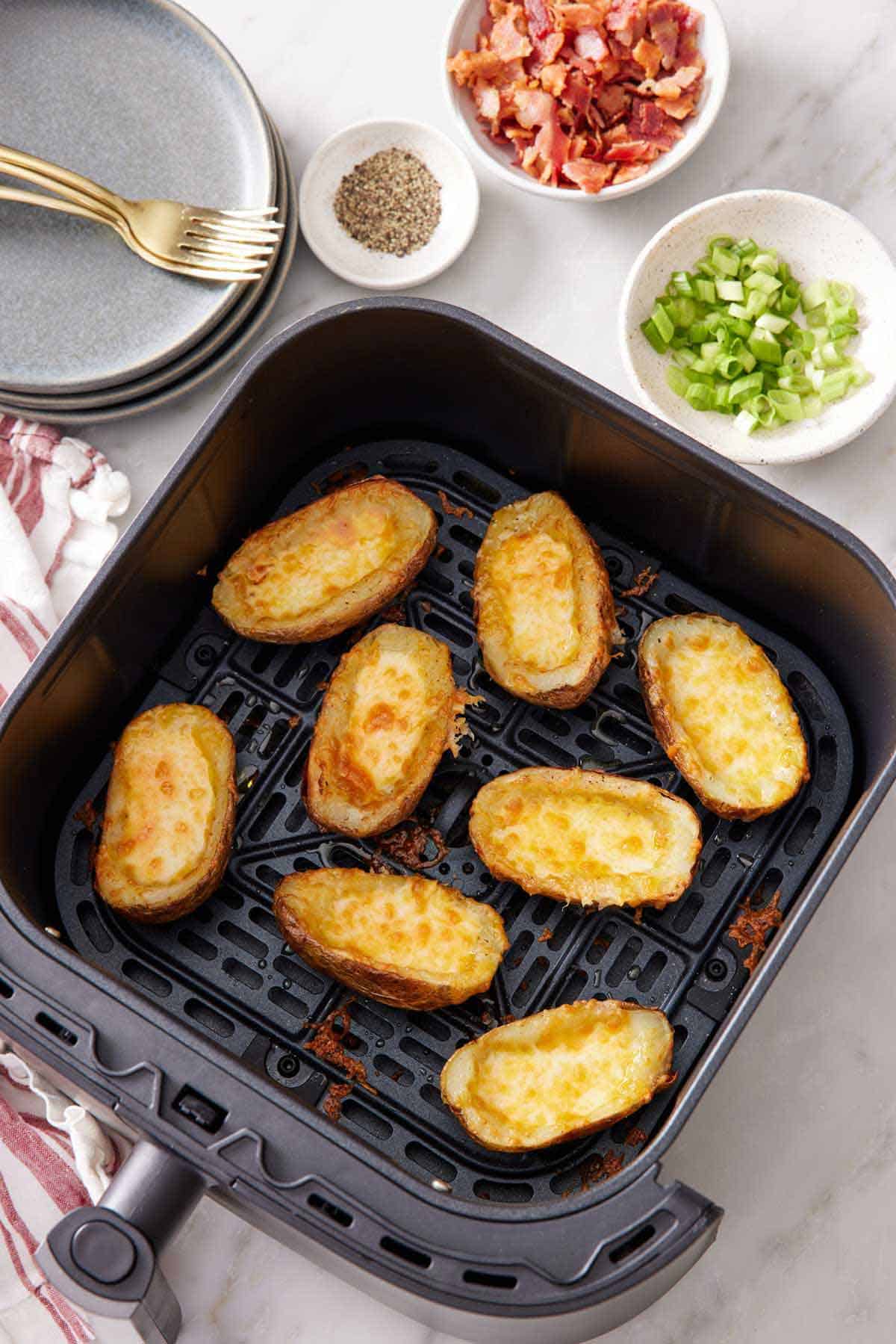 Overhead view of an air fryer basket with potato skins. Green onions, bacon, pepper, plates, and forks on the side.