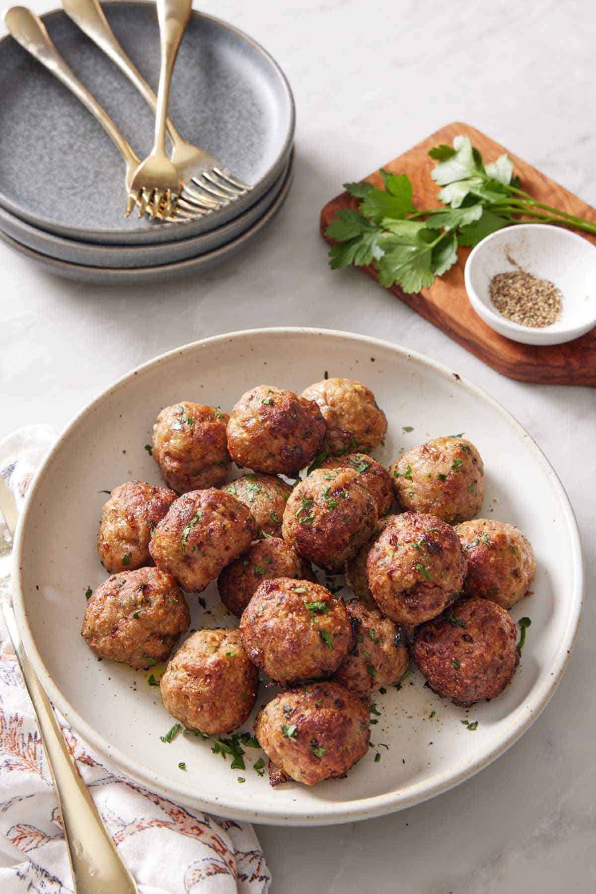 A bowl of air fryer turkey meatballs garnished with chopped parsley. A stack of plates, forks, some parsley, and bowl of pepper in the background.