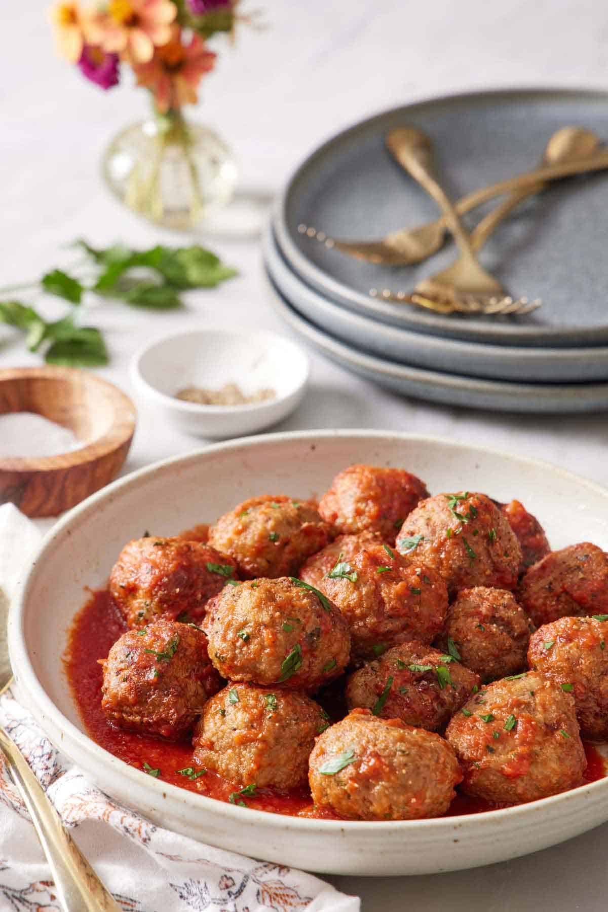 A bowl of air fryer turkey meatballs coated in tomato sauce. A stack of plates in the back with forks, salt and pepper, and parsley.