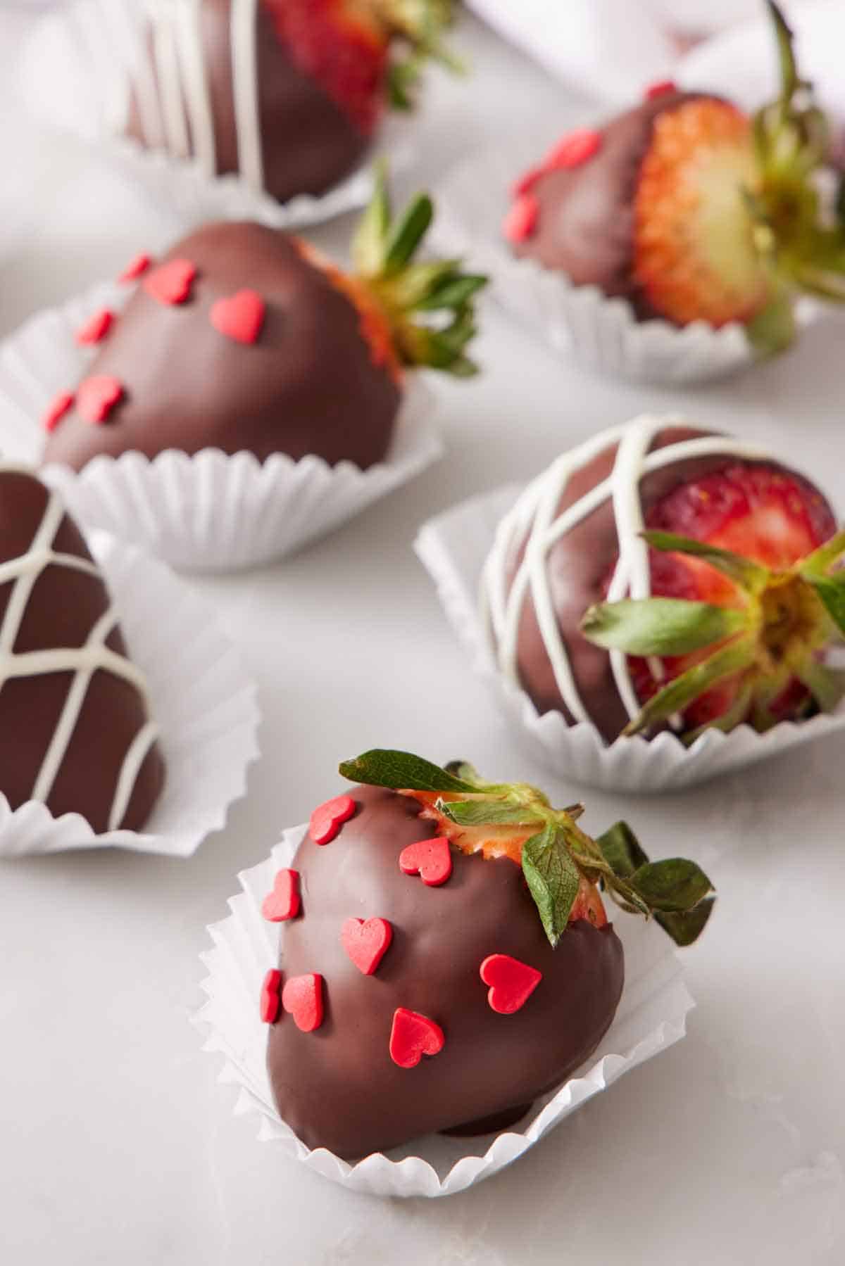 Multiple chocolate covered strawberries in paper liners, some with heart sprinkles and some with white chocolate drizzle.